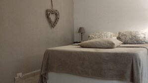 Une chambre à coucher avec un lit, une table de chevet, un décor mural en forme de cœur, un petit tabouret en bois et un sol carrelé.