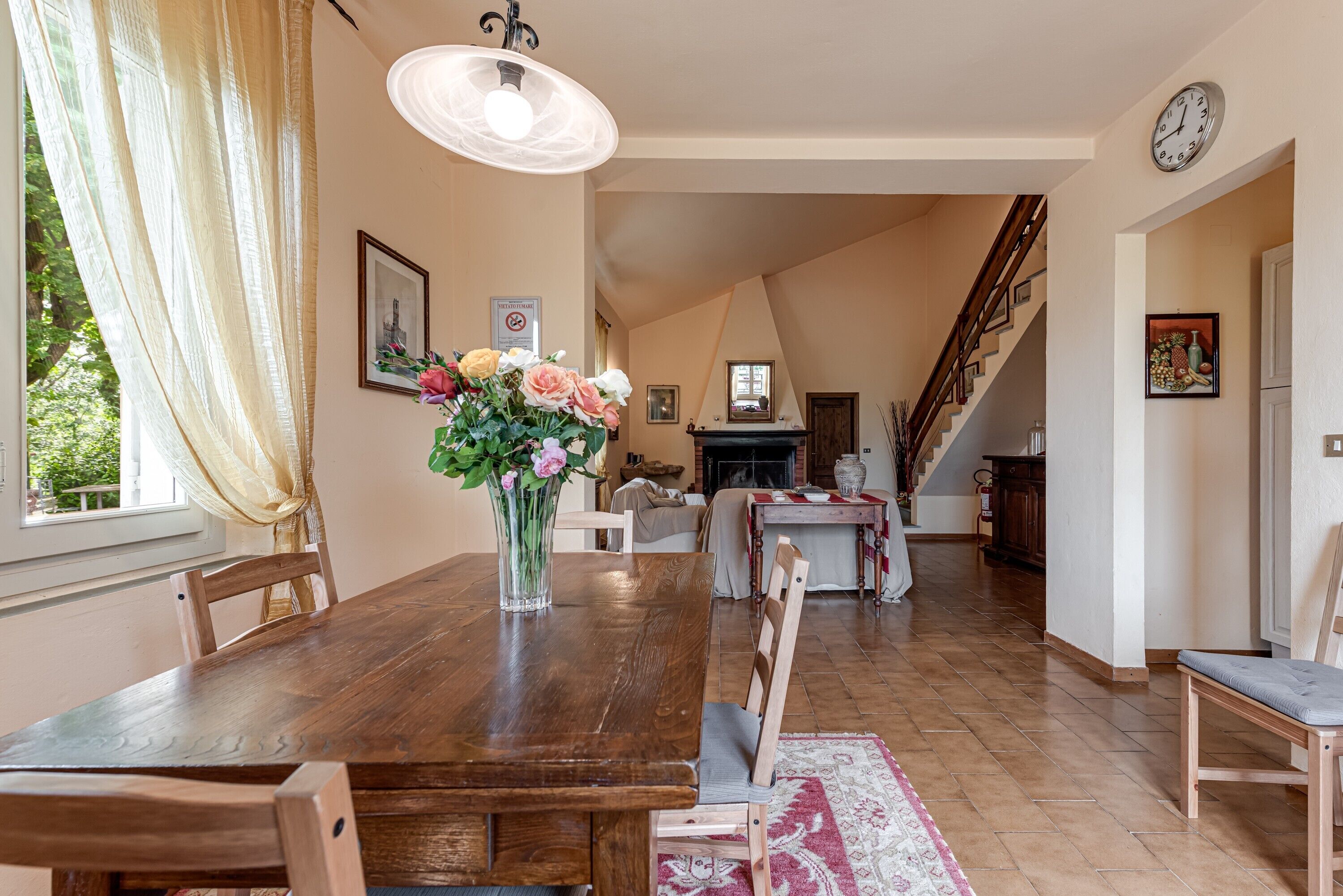 A dining area with a wooden table, chairs, a vase with flowers, a chandelier, and a staircase leading to another room.