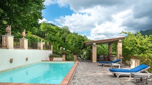 Ein Schwimmbad mit einer Steinterrasse, Liegestühlen und einer Pergola.