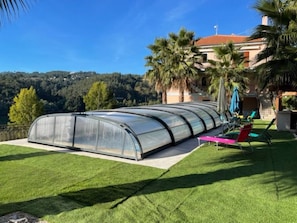A greenhouse structure on a lawn with a view of a hill and a building in the background.
