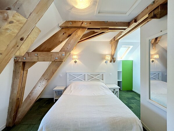 Un dormitorio con techo inclinado, vigas de madera, cama, mesitas de noche, lámparas de pared y una puerta verde.