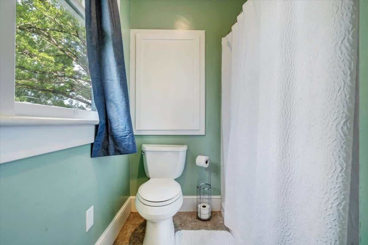 A bathroom with a toilet, a white towel hanging on the wall, and a window with a curtain.