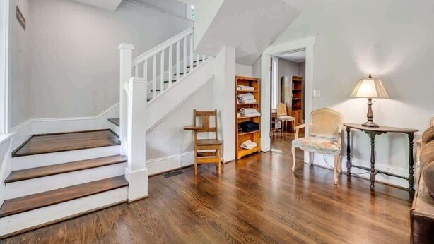 Una escalera con barandillas blancas que conduce a una plataforma, piso de madera, una silla, una mesita con una lámpara y una estantería.