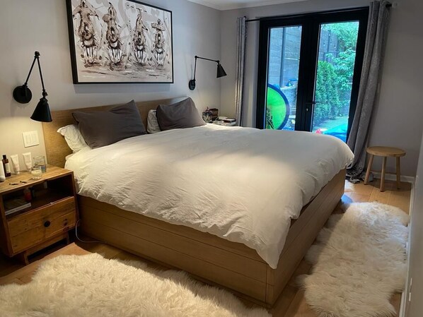 A bedroom with a large bed, a nightstand, a lamp, a painting on the wall, and a view of greenery outside.