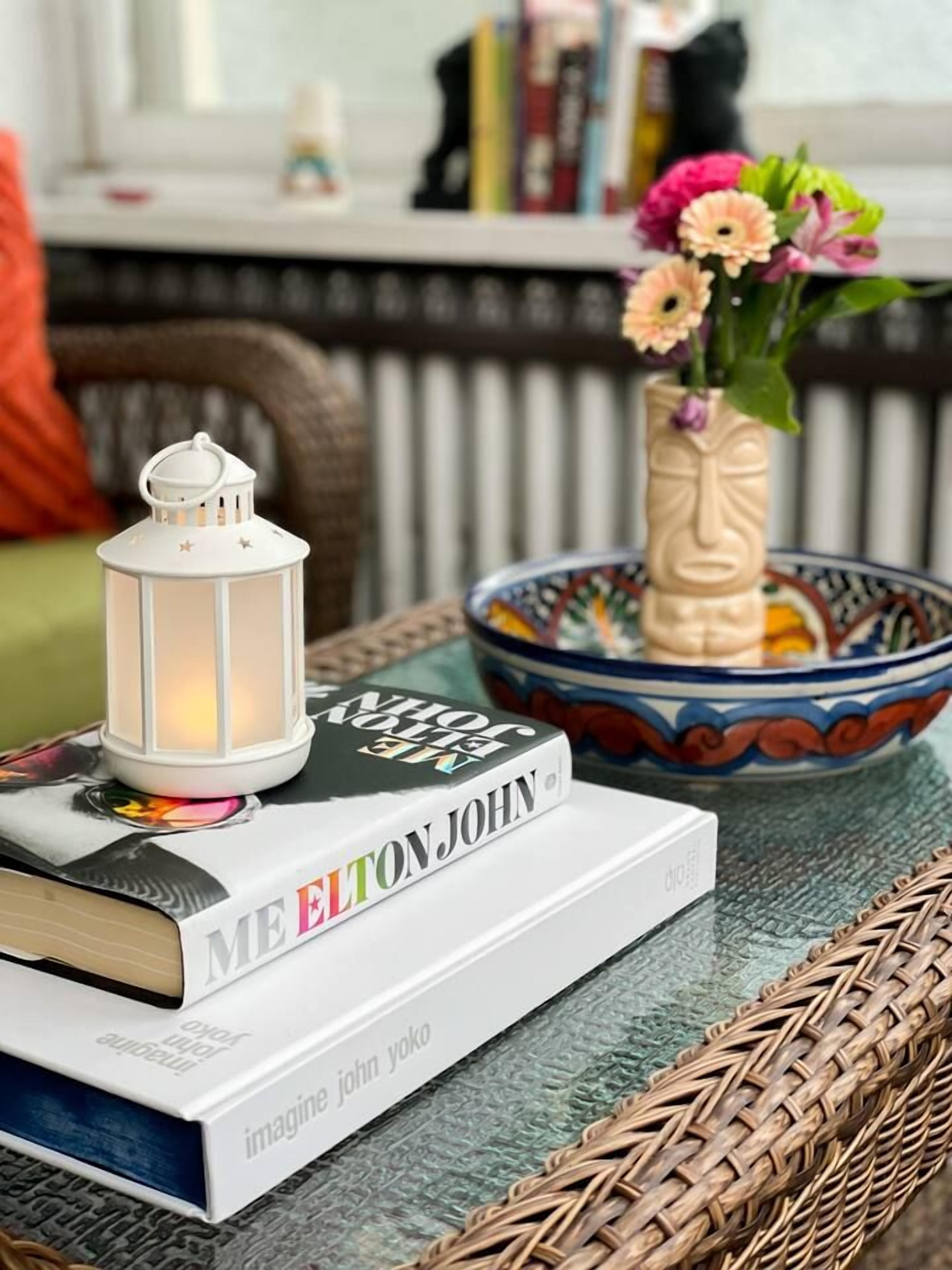 Un livre intitulé « MEELTON JOHN » est posé sur une table en osier, à côté d’une lanterne blanche et d’un vase décoratif rempli de fleurs.