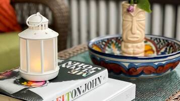 Un livre intitulé « MEELTON JOHN » est posé sur une table en osier, à côté d’une lanterne blanche et d’un vase décoratif rempli de fleurs.