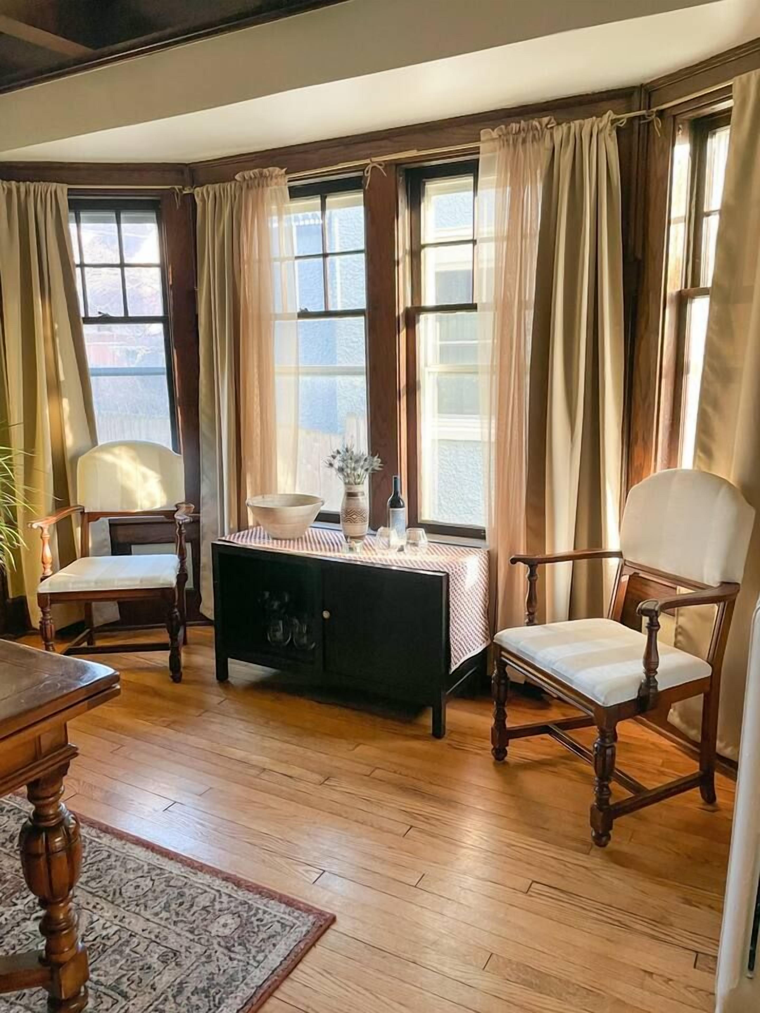 Una habitación con suelo de madera, dos sillones tapizados, una mesa de madera, un armario negro y una ventana con cortinas.