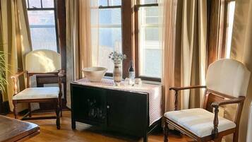 Una habitación con suelo de madera, dos sillones tapizados, una mesa de madera, un armario negro y una ventana con cortinas.