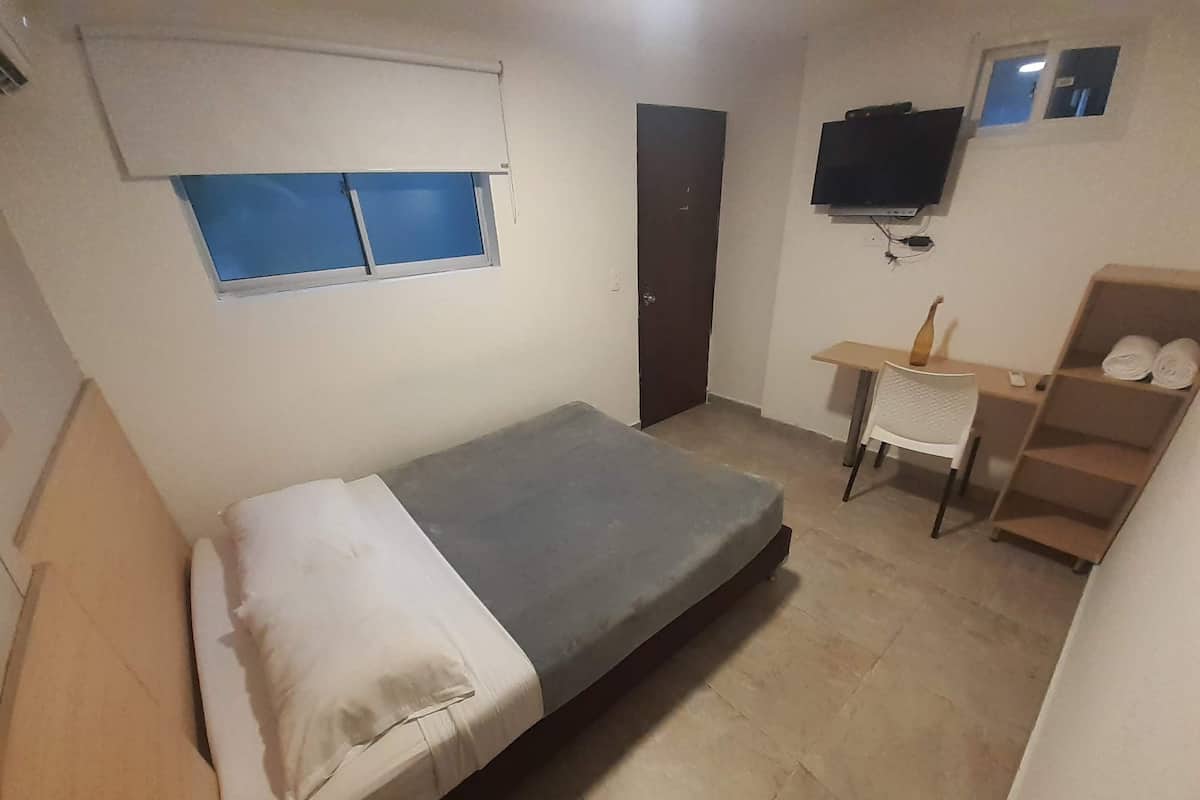 Quarto pequeno e compacto com uma cama, escrivaninha, cadeira, TV fixada na parede e janela com vidro azulado.