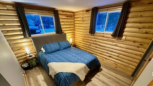 A bedroom with a wooden log wall, a bed with blue bedding, a nightstand with a lamp, and windows with curtains. - Waterfront Log Home with Beach and Hot Tub. Pet Friendly! (Clandeboye)