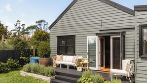 Une maison moderne à l’extérieur gris, avec un porche abrité comprenant un canapé blanc et une petite table, et une porte-fenêtre coulissante donnant sur un patio.