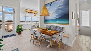 A dining area with a wooden table, wicker chairs, and a large ocean view painting. - Call Me The Breeze - Private Home At The Shores 3 Bedroom Home by RedAwning (South Padre Island)
