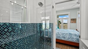 A modern bathroom with a glass shower enclosure, a visible showerhead, and a large window with blinds. - Call Me The Breeze - Private Home At The Shores 3 Bedroom Home by RedAwning (South Padre Island)