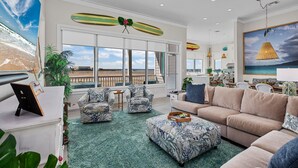 A spacious living room with a large window overlooking the beach, a ceiling fan, and a surfboard decoration. - Call Me The Breeze - Private Home At The Shores 3 Bedroom Home by RedAwning (South Padre Island)