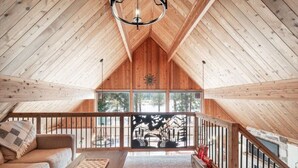 A cozy attic room with a wooden ceiling, a sofa, a table, and a view of the outdoors through large windows. - Waterfront Loon Cottage | Private Boat Dock | Hot Tub (Island Park)