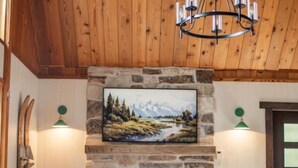 A cozy living room with a stone fireplace, a painting above it, and a plaid sofa. - Waterfront Loon Cottage | Private Boat Dock | Hot Tub (Island Park)