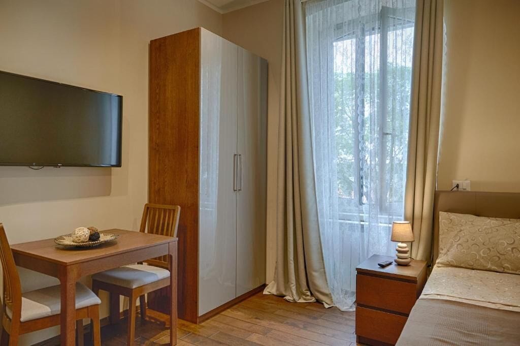 Une chambre comprenant un lit, un téléviseur fixé au mur, une table en bois avec des chaises, une armoire et une fenêtre avec des rideaux.