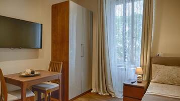 A room with a bed, a TV mounted on the wall, a wooden table with chairs, a wardrobe, and a window with curtains.