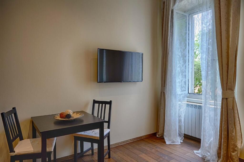 A room with a wooden table and chairs, a flat-screen TV mounted on the wall, and a window with sheer curtains.
