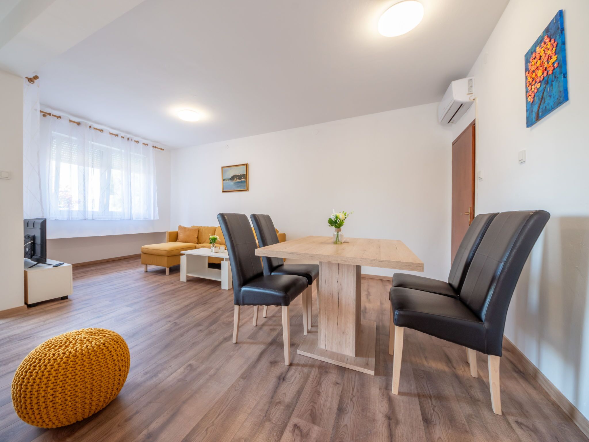 A modern living room with a wooden dining table, black leather chairs, a yellow ottoman, a sofa, and a TV.