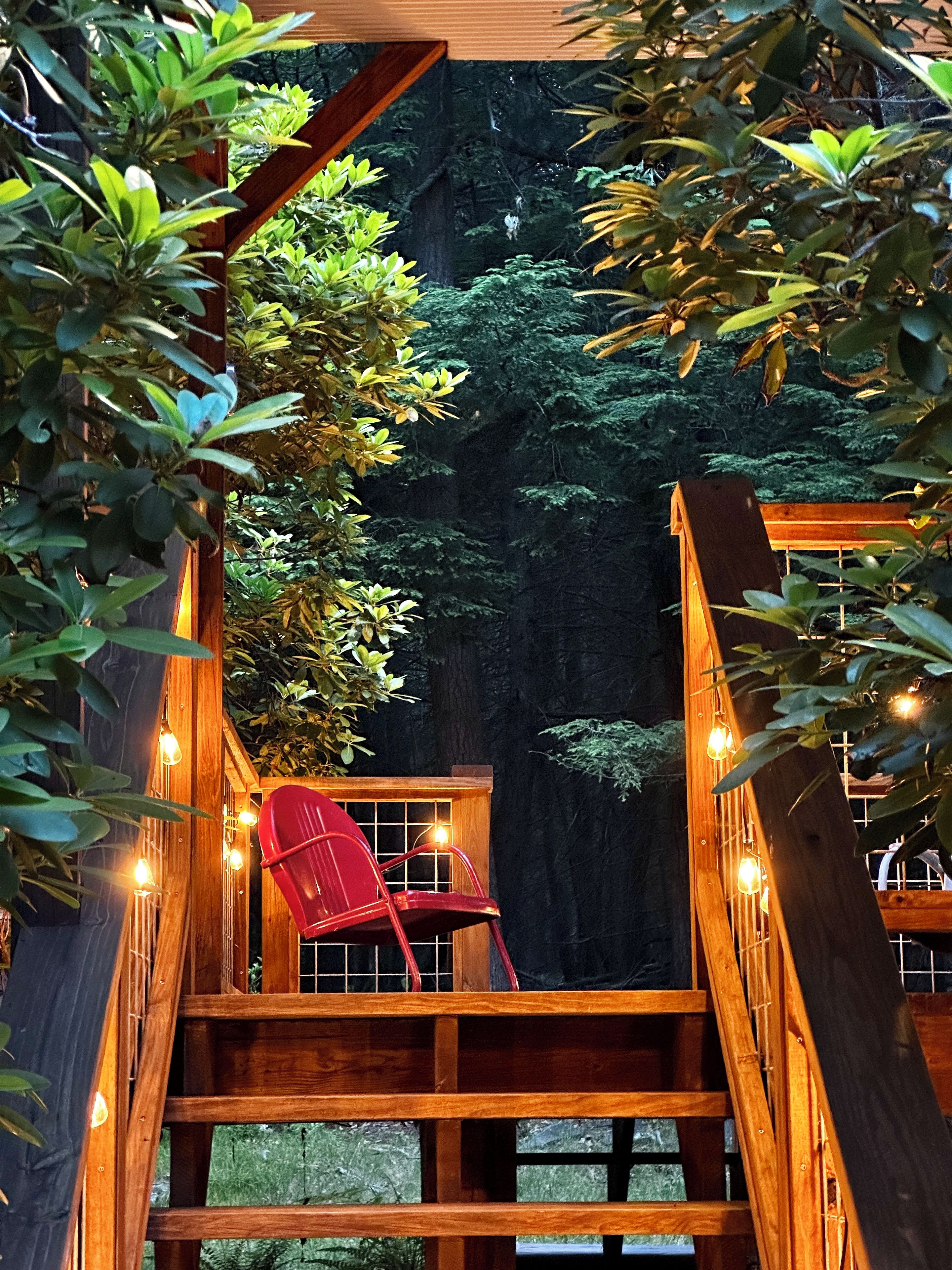 A wooden deck with a red chair, surrounded by greenery and string lights.