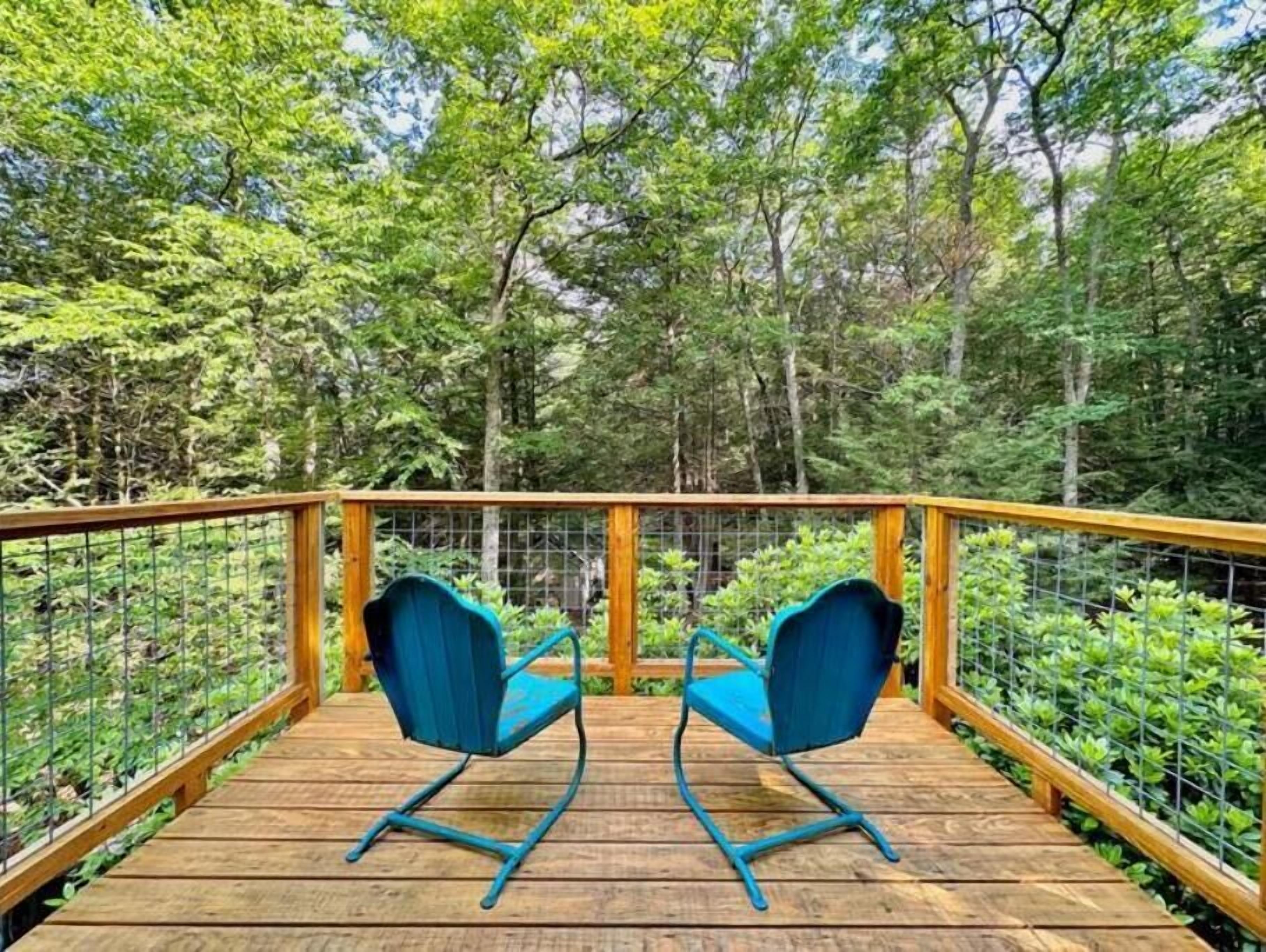 A wooden deck with two blue chairs and a metal railing, surrounded by dense green foliage.