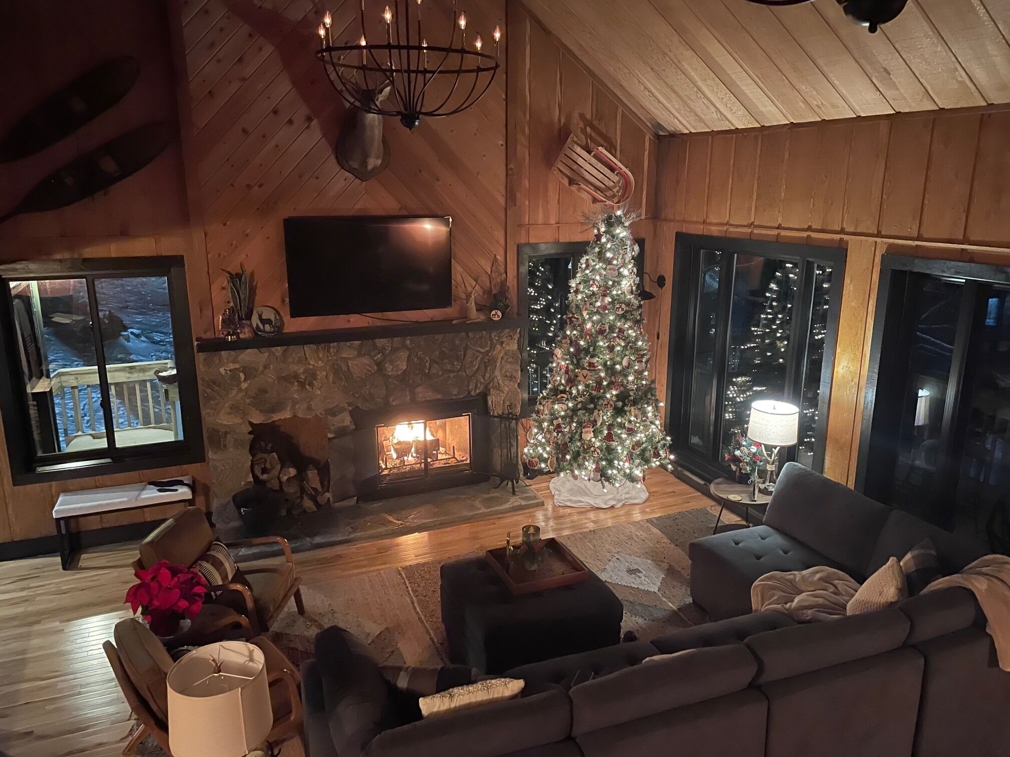 A cozy living room with a stone fireplace, a Christmas tree, and a large window overlooking a deck.