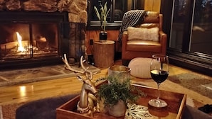 A cozy living room with a stone fireplace, a leather armchair, a wooden coffee table with a glass of wine, and a deer figurine.
