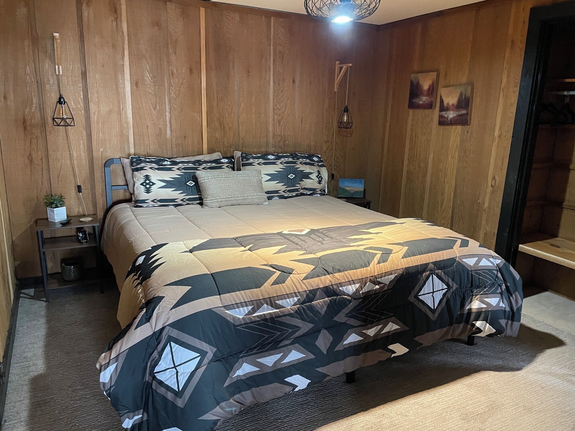 A bedroom with a wooden wall, a bed with patterned bedding, a nightstand with a plant, and a hanging light fixture.