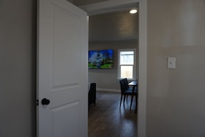 A room with a white door, a dining table with chairs, a painting on the wall, and a window with blinds.
