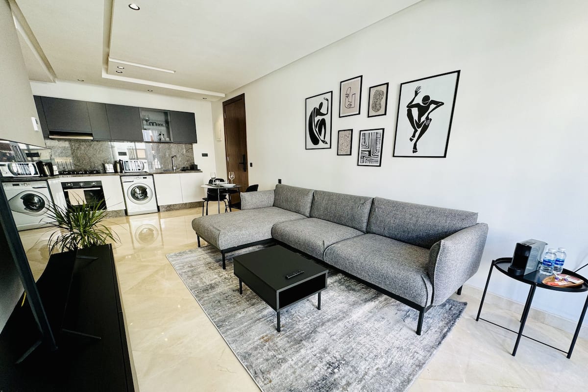 A modern living room with a grey sectional sofa, a black coffee table, and a grey rug. There is a dining area with a small table and chairs, and a kitchen with a built-in oven and washing machine.