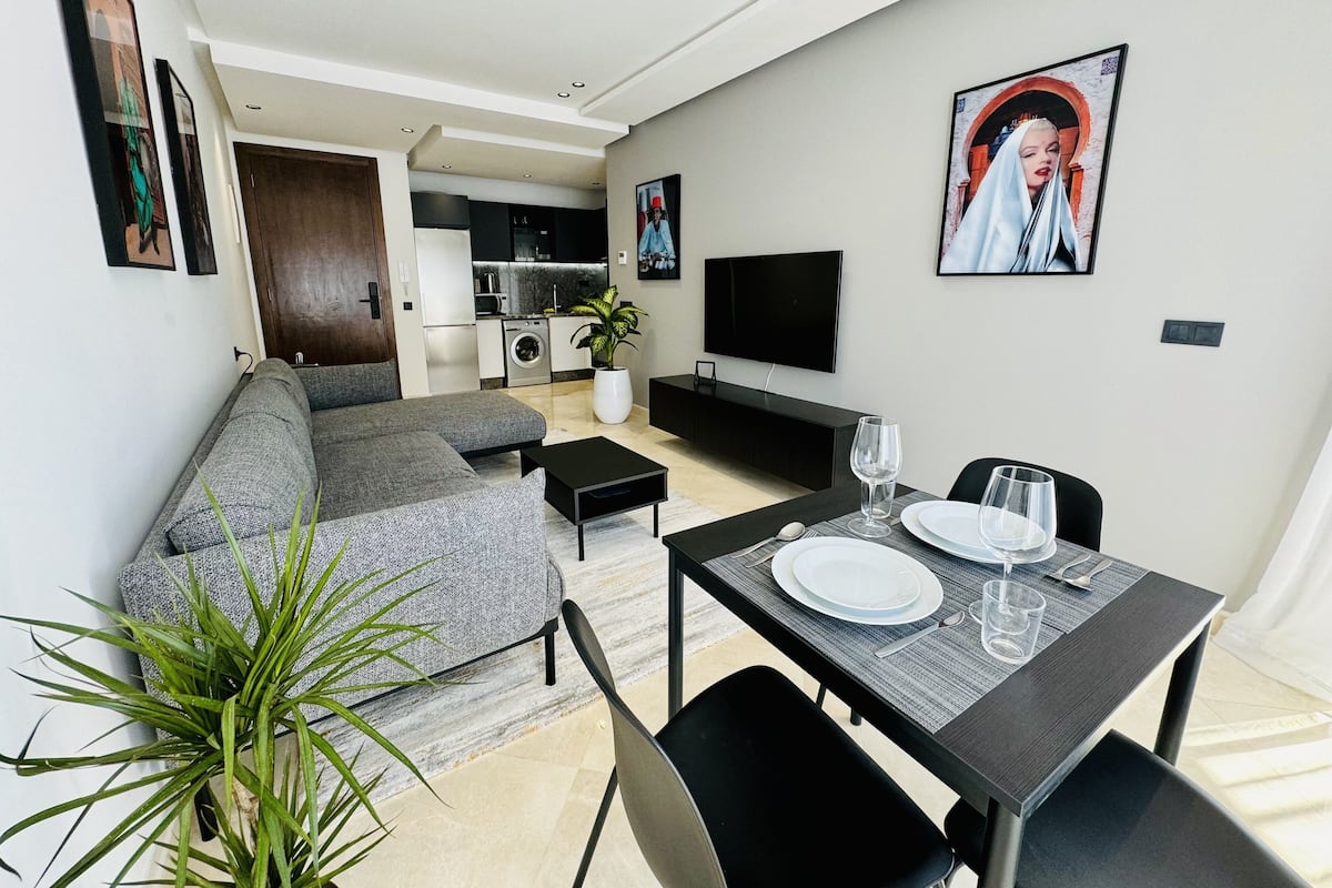 A modern living room with a grey sofa, a dining table set for two, a flat-screen TV, and a kitchen in the background.