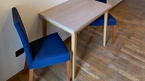A small dining area with a wooden table and two blue chairs. A framed picture hangs on the wall above the table.