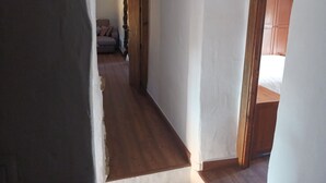 Un couloir étroit, doté d’un plancher en bois et de murs blancs, mène à une pièce où se trouvent un lit et des meubles en bois.