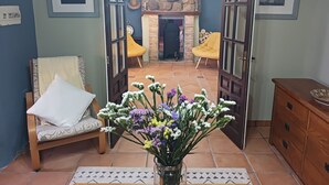 Une porte ouverte donne sur un salon agrémenté d’une cheminée en pierre, d’un fauteuil jaune et d’une armoire en bois.