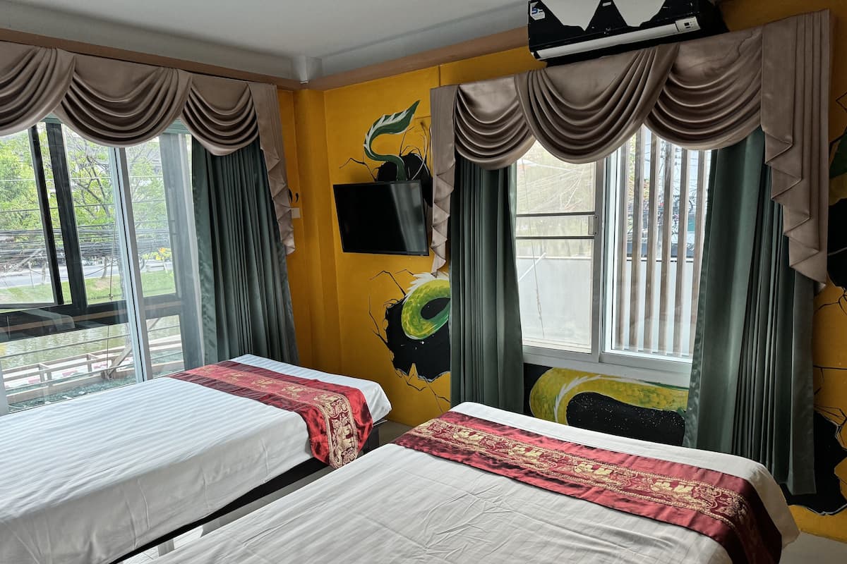 A room with two beds, a TV mounted on the wall, and large windows with curtains.