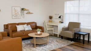 Una sala de estar con un sofá de cuero marrón, un sillón beige, una mesa de centro de madera y una alfombra estampada. Hay un escritorio blanco con una silla, un cuadro enmarcado en la pared y una ventana con persianas.