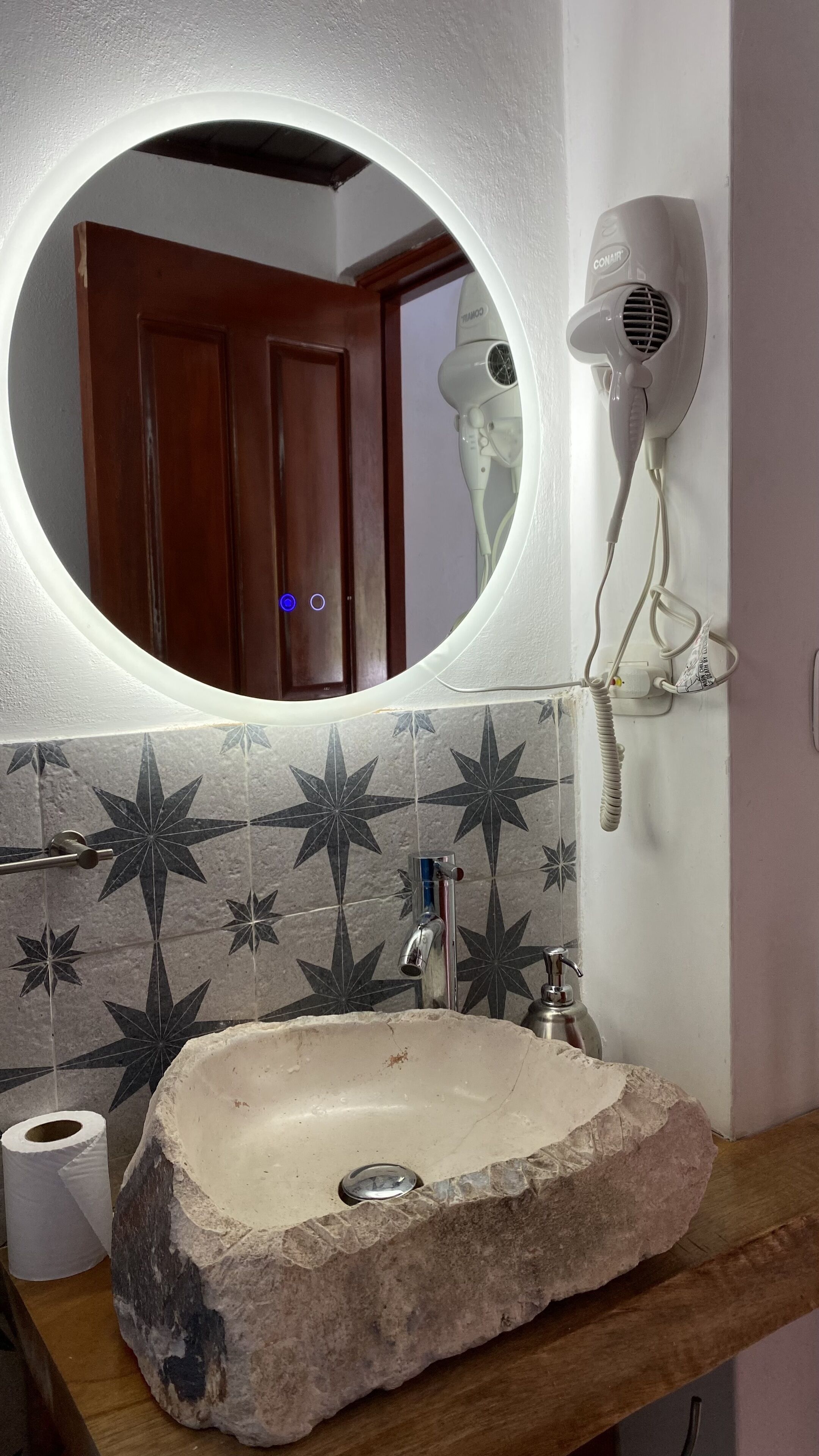 Un baño con lavamanos de piedra, un espejo con luz, un secador de pelo y un rollo de papel higiénico.