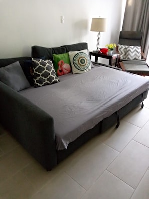 Ein modernes Wohnzimmer mit einem grauen Sofa, gemusterten Kissen, einem kleinen Beistelltisch mit einer Lampe und Blick ins Freie durch ein Fenster.