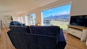 A living room with a dark blue sofa, wooden flooring, a television, and large windows offering a mountain view. - Aonach View near Fort William (Highland Council)