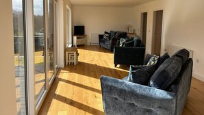 A spacious living area with a large sliding glass door leading to an outdoor patio, a dark grey sofa set, and a small white side table with a television. - Aonach View near Fort William (Highland Council)