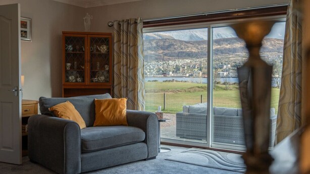 A living room with a grey sofa, a wooden cabinet, and a view of a town and mountains through a large glass door. - Ben View located in Teslaig, Fort Willam (Treslaig)