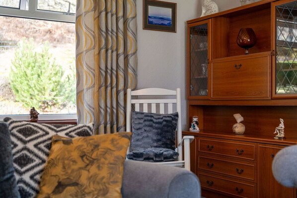 A cozy living room with a white chair, a wooden cabinet with glass doors, and patterned cushions.