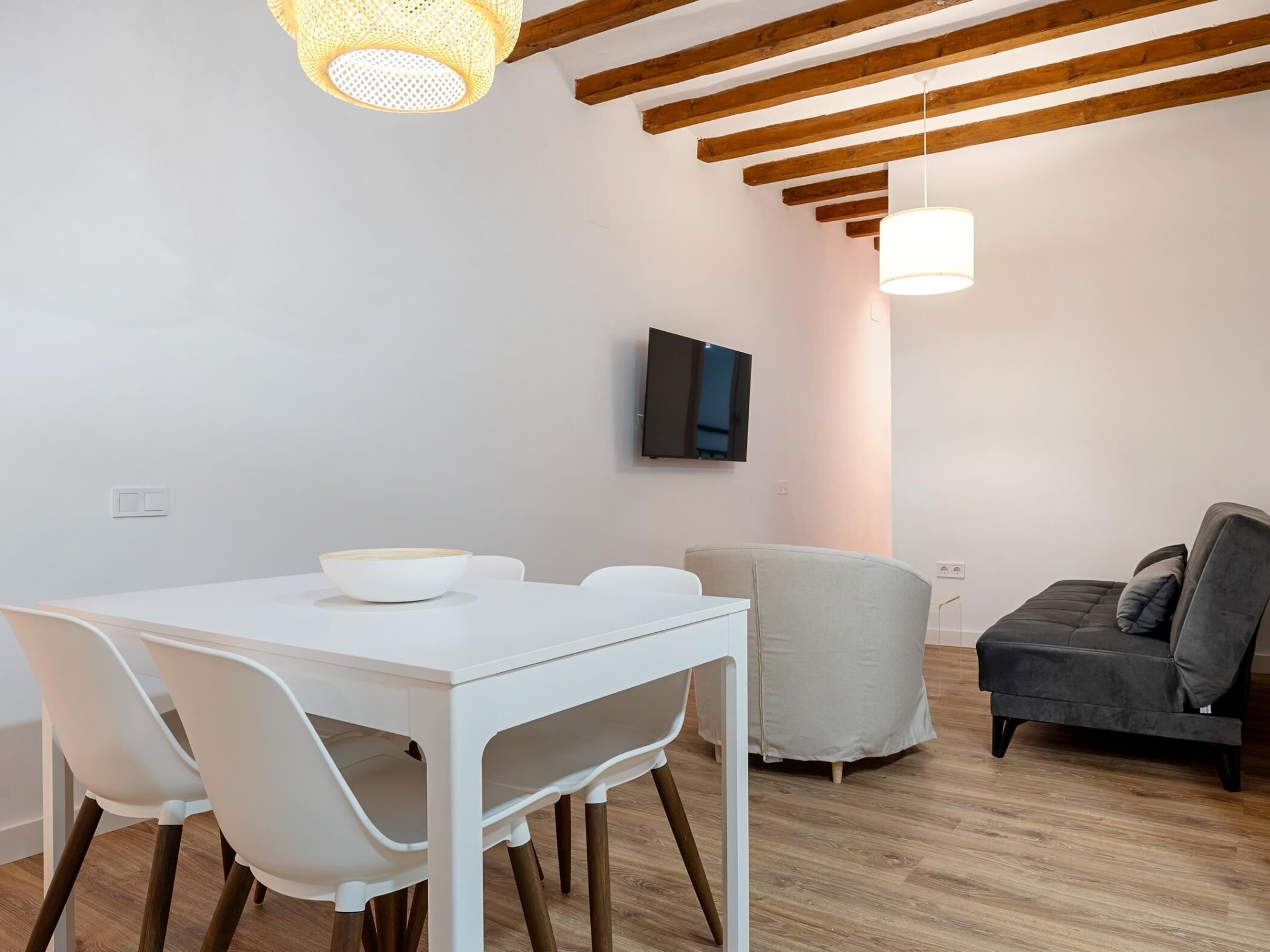 Un comedor moderno con una mesa y sillas blancas, un sofá gris, vigas de madera en el techo y un televisor montado.