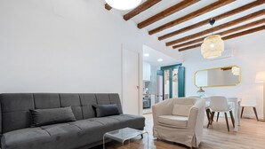 Ein modernes Wohnzimmer mit einem grauen Sofa, einem transparenten Couchtisch, einem beige farbenen Sessel und einem Essbereich mit weißem Tisch und Stühlen.
