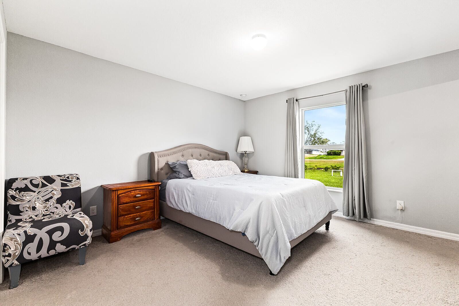 A bedroom with a bed, nightstand, dresser, and a view of a green landscape through a window.