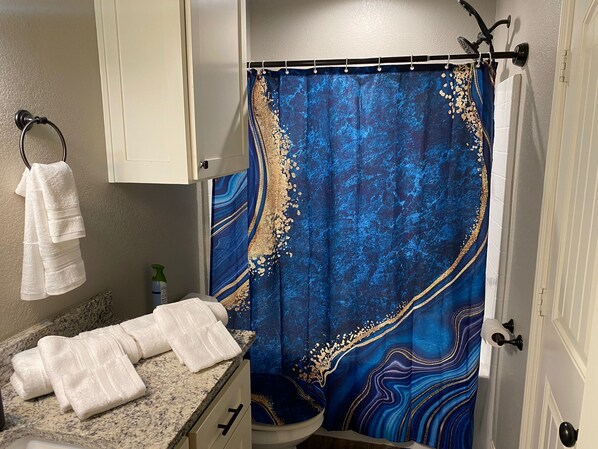 Une salle de bain avec un rideau de douche bleu, un porte-serviettes avec des serviettes blanches, une bouteille de shampoing et un comptoir en granit.