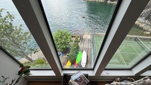 Une pièce dotée d’une grande fenêtre donnant sur une étendue d’eau, un court de tennis et un espace vert avec des arbres et un banc.