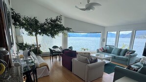 Un salon spacieux doté d’une grande fenêtre offrant une vue sur un lac et des montagnes. On y trouve plusieurs coins salon, dont un canapé et une table à manger avec des chaises.