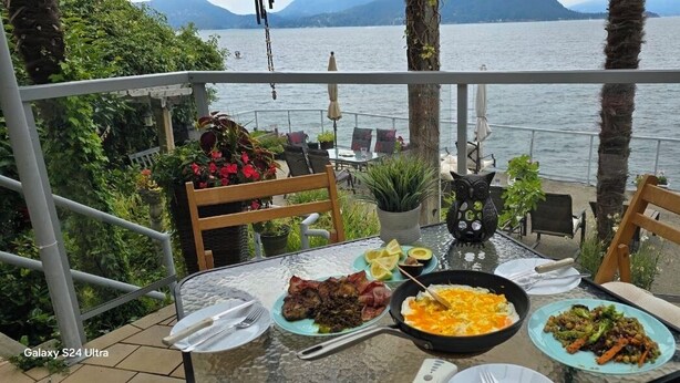 Une table à manger avec des assiettes remplies de nourriture, une poêle noire et une statuette de hibou décorative sur un balcon donnant sur une étendue d’eau.